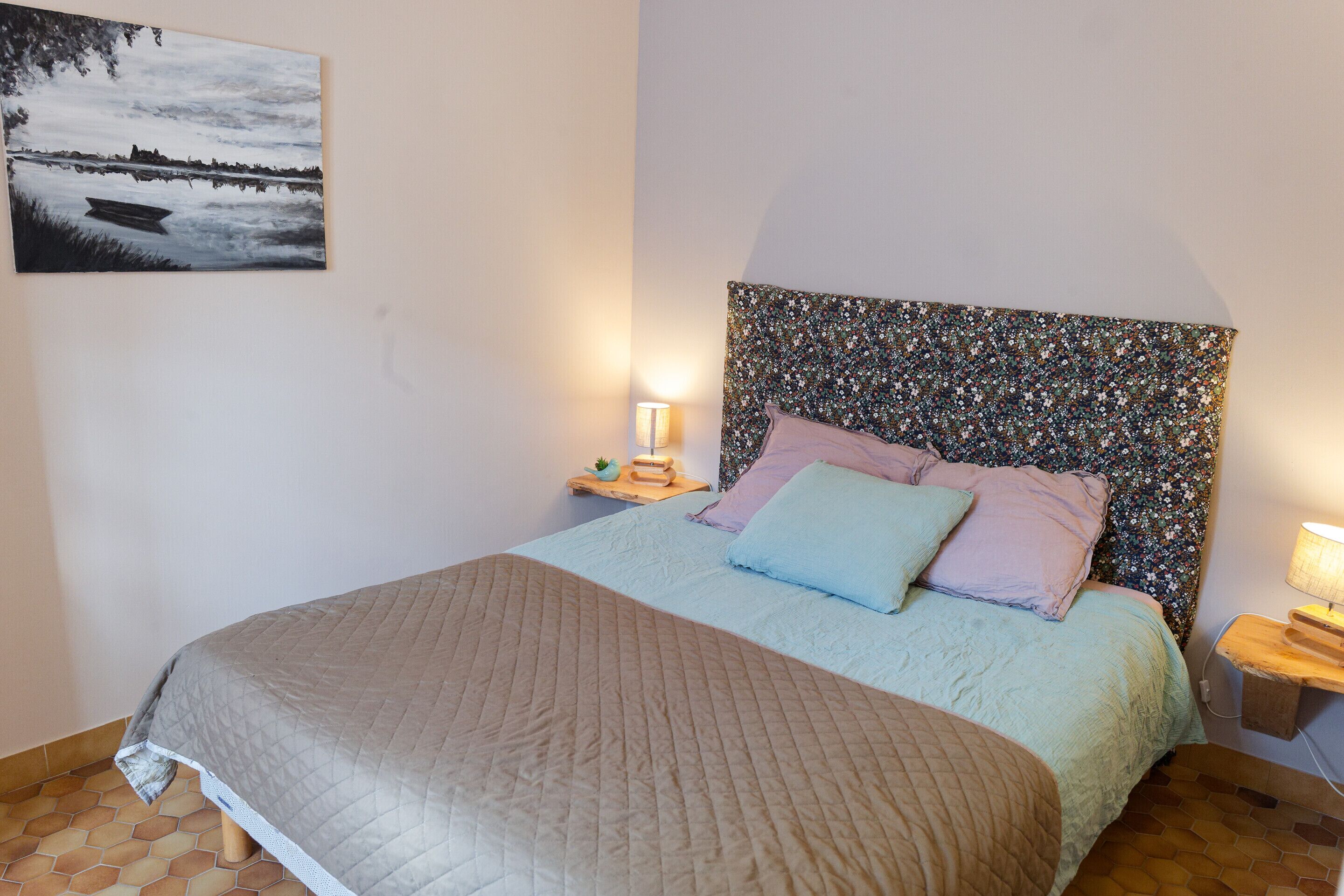 A bedroom with a bed, a headboard, a nightstand, a lamp, and a painting on the wall.