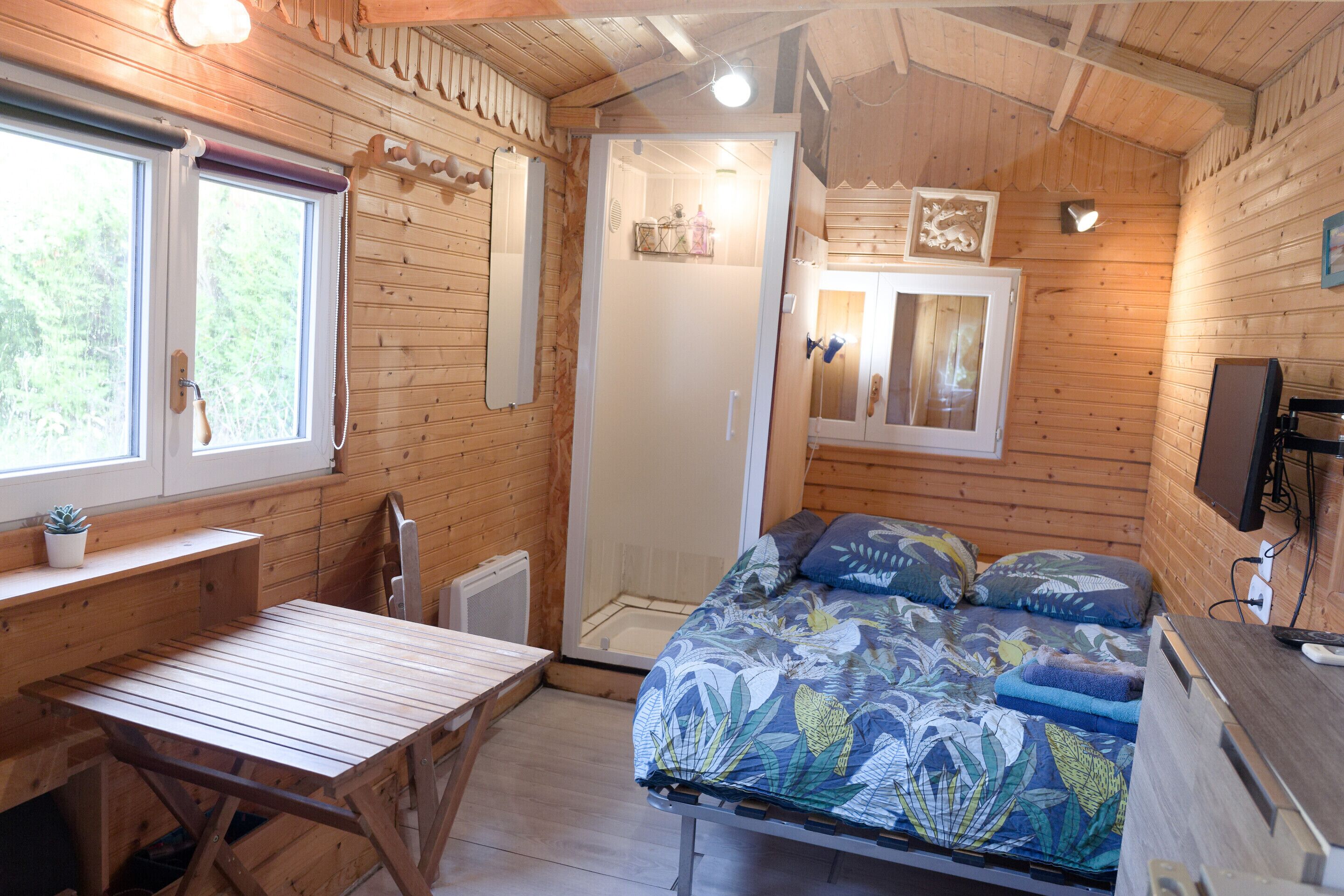 A cozy wooden cabin with a bed, a desk, and a shower area.
