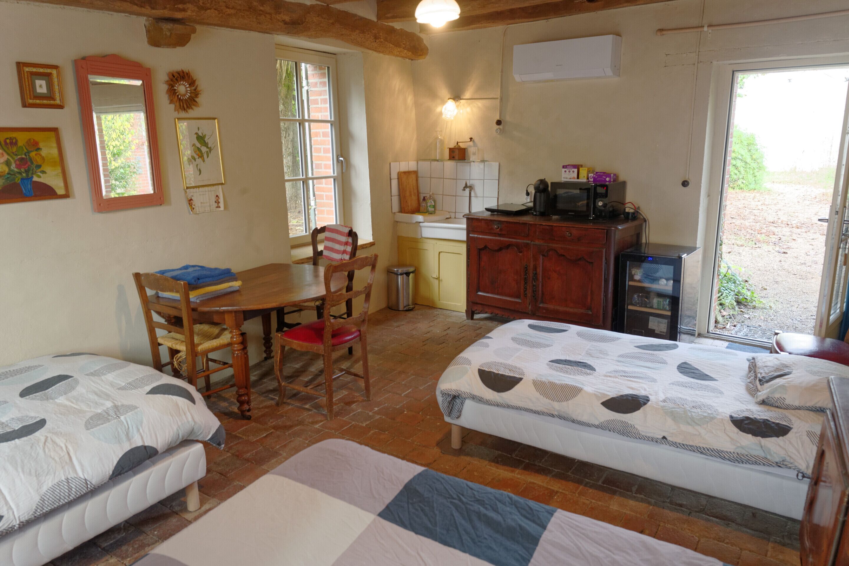 A cozy room with two beds, a wooden dining table, chairs, a small kitchen area with a microwave, and a window with a view of the outdoors.