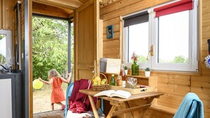 A wooden cabin interior with a dining area, a table with chairs, a window with a red blind, and a door leading outside.