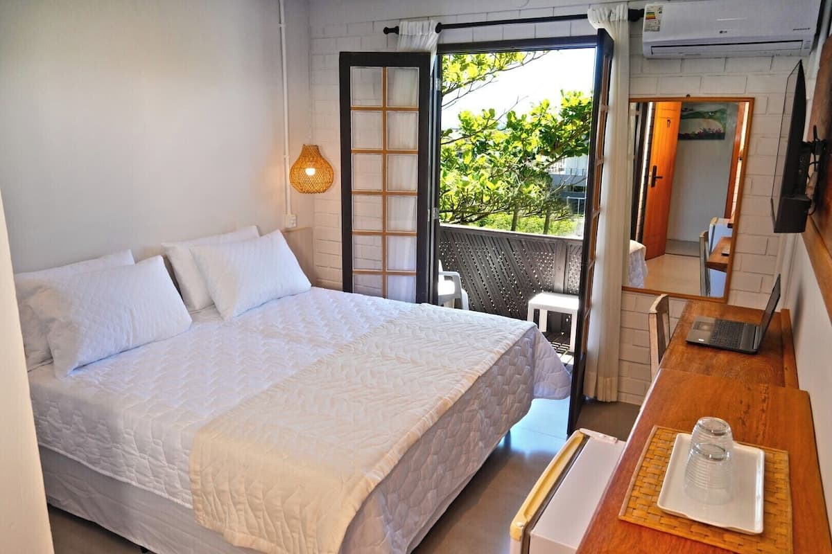 Ein Schlafzimmer mit einem großen Bett, einem Schreibtisch mit einem Laptop, einem Fenster mit Blick auf Grünflächen und einer Schiebetür zu einem Außenbereich.