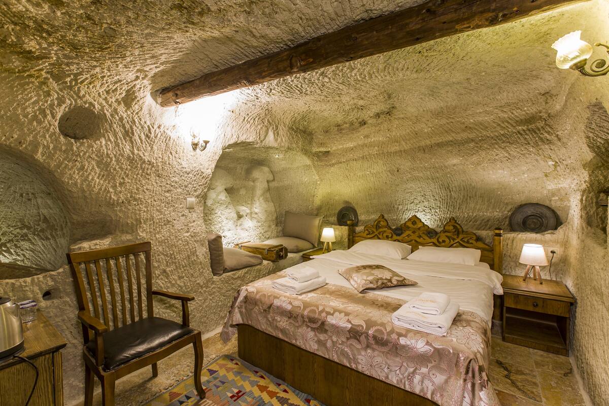 Una habitació rústica de pedra amb un llit de fusta, una cadira i una tauleta de nit amb una làmpada.