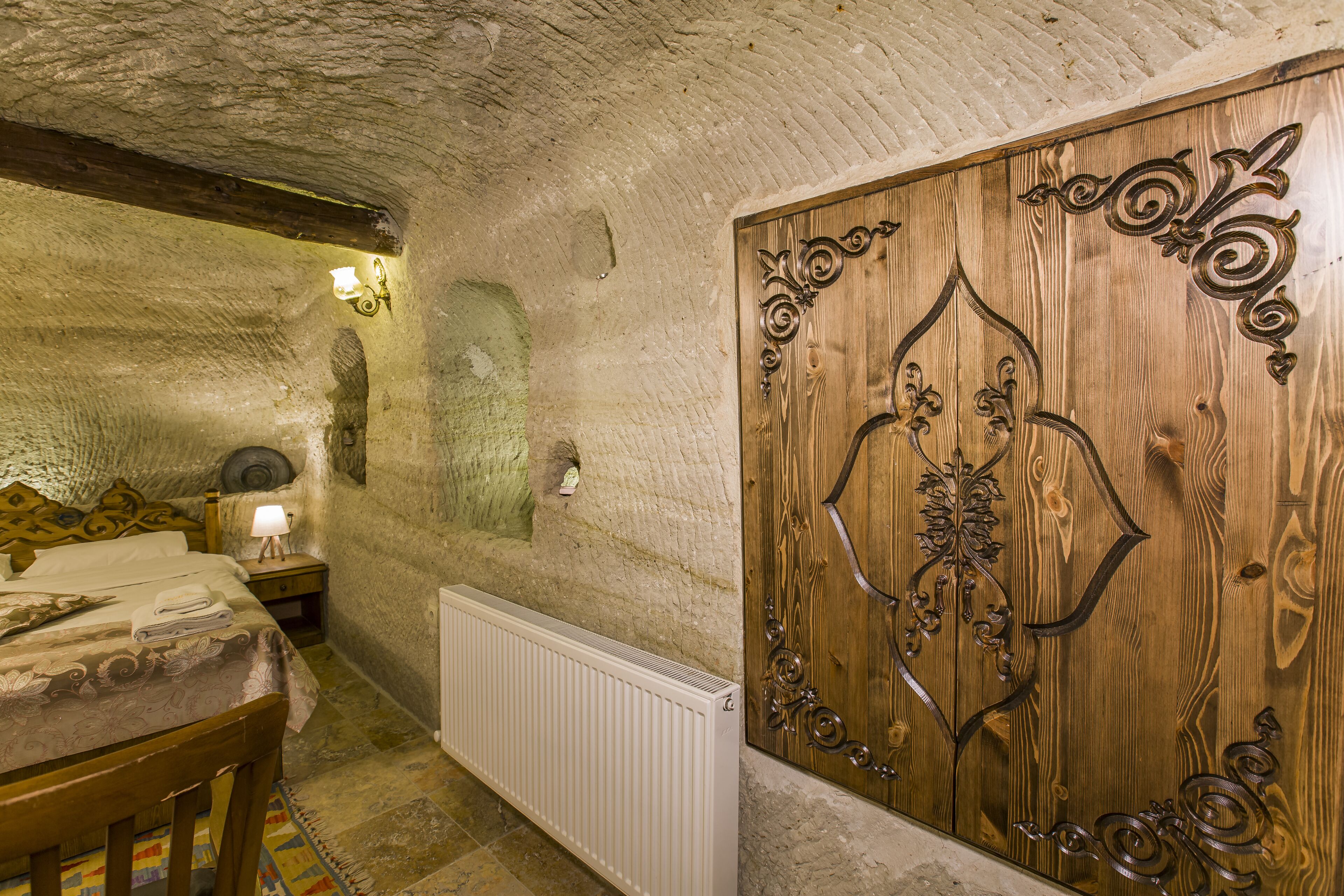 A rustic room with a wooden door, stone walls, a bed with a patterned bedspread, a nightstand with a lamp, and a radiator.