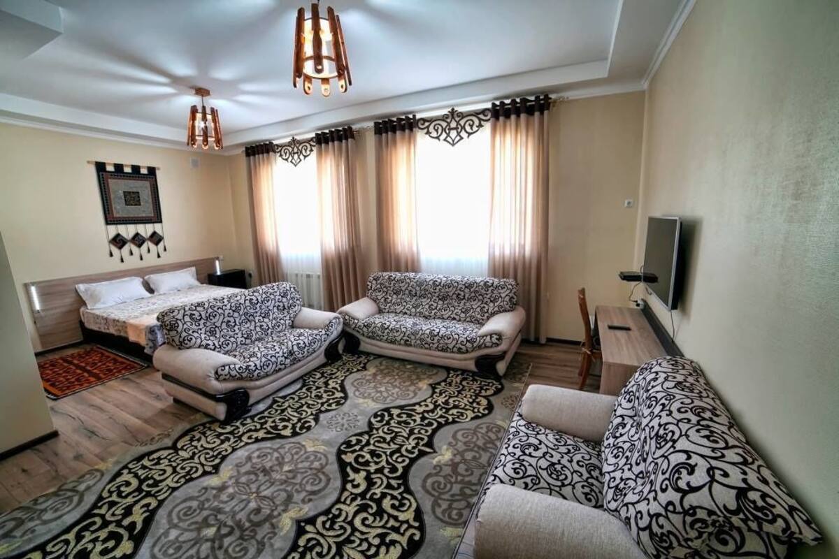 A modern hotel room with a bed, two patterned armchairs, a sofa, a TV, and a large window with curtains.