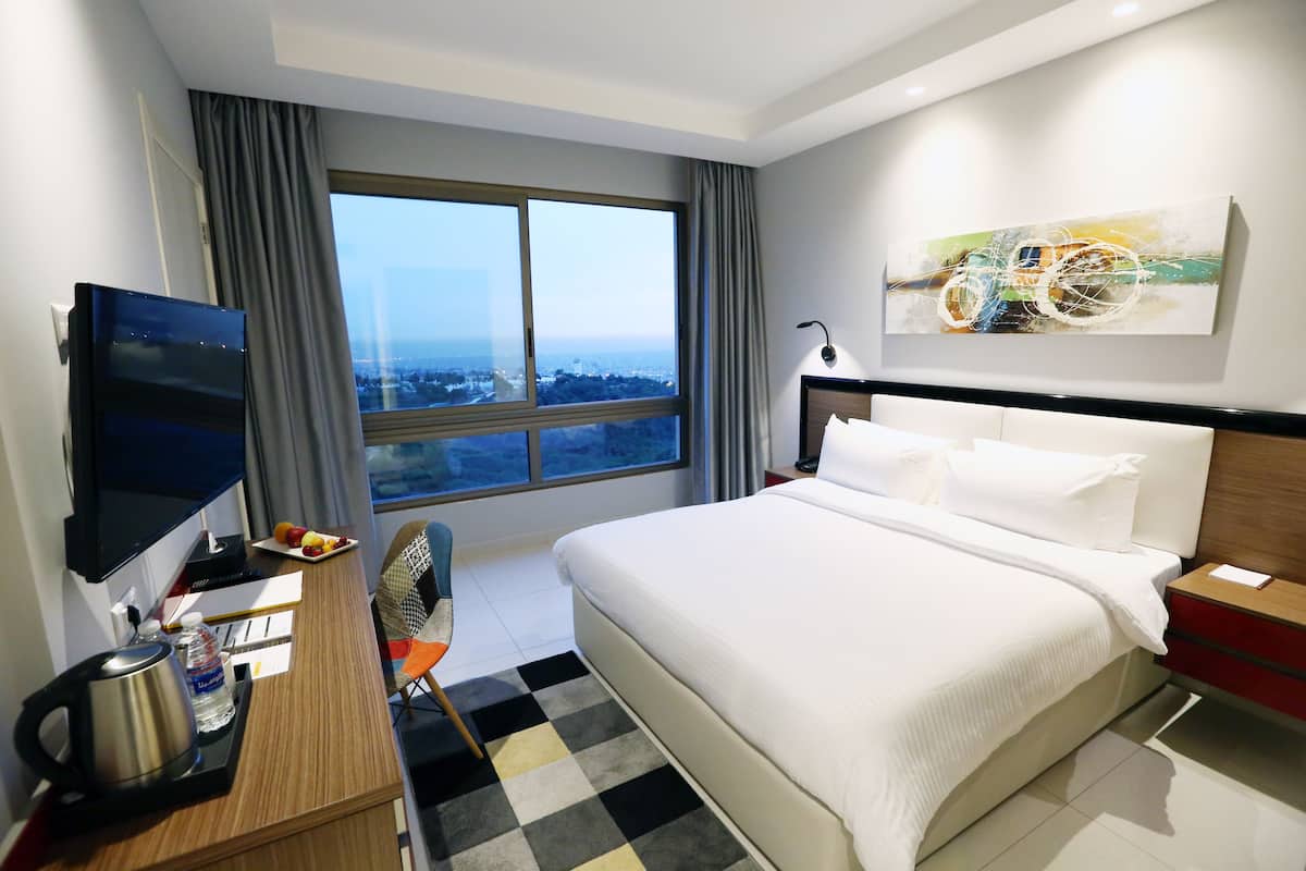Una camera d'hotel con un grande letto, una TV a schermo piatto, una scrivania con bollitore e bottiglia d'acqua, una poltrona e vista sulla città attraverso un'ampia finestra.