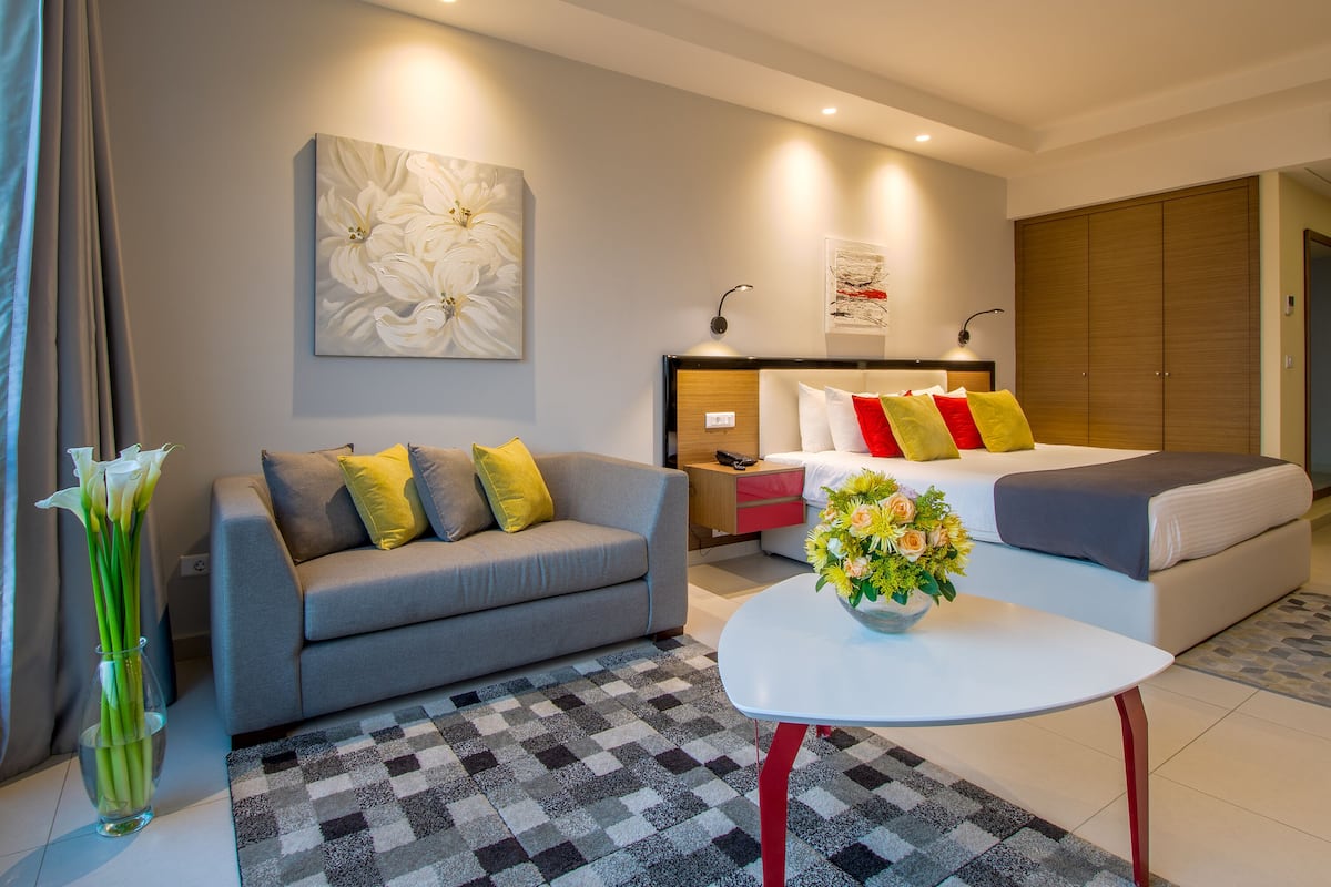 Una camera d'albergo moderna con un divano grigio, un letto bianco con cuscini rossi e gialli, un piccolo tavolino bianco e un grande quadro astratto appeso alla parete.
