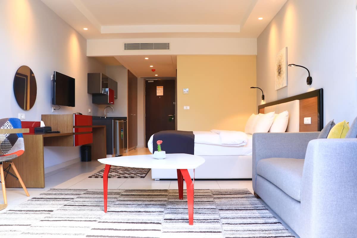 Un soggiorno moderno con un divano grigio, un tavolino da caffè bianco e una poltrona rossa. È presente un angolo cottura con frigorifero, microonde e pensili. Una TV è montata a parete, e una porta conduce in un'altra stanza.