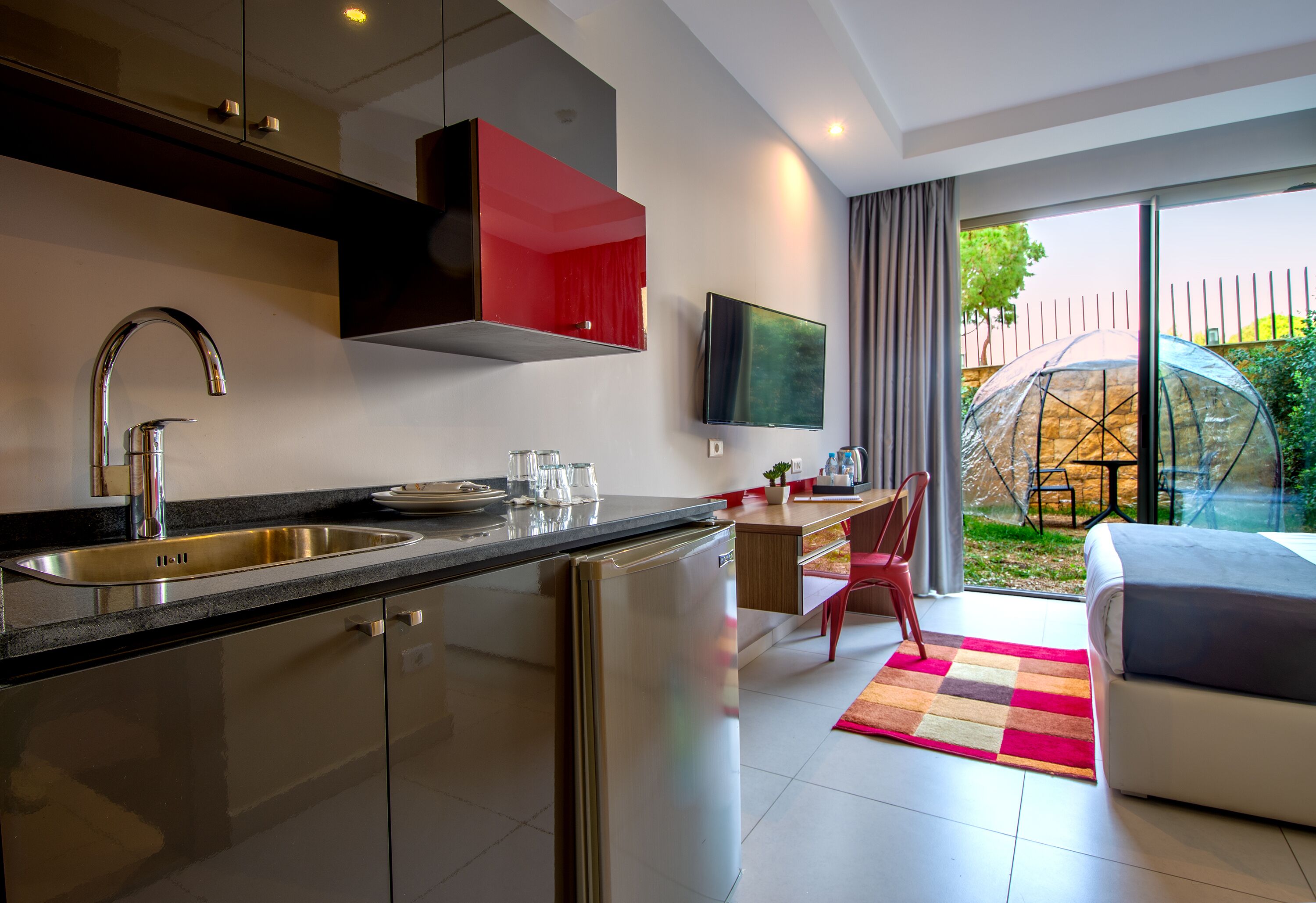 A modern kitchen with a stainless steel sink and a built-in refrigerator. There is a dining area with a table and chairs, and a large window offering a view of the outdoors.
