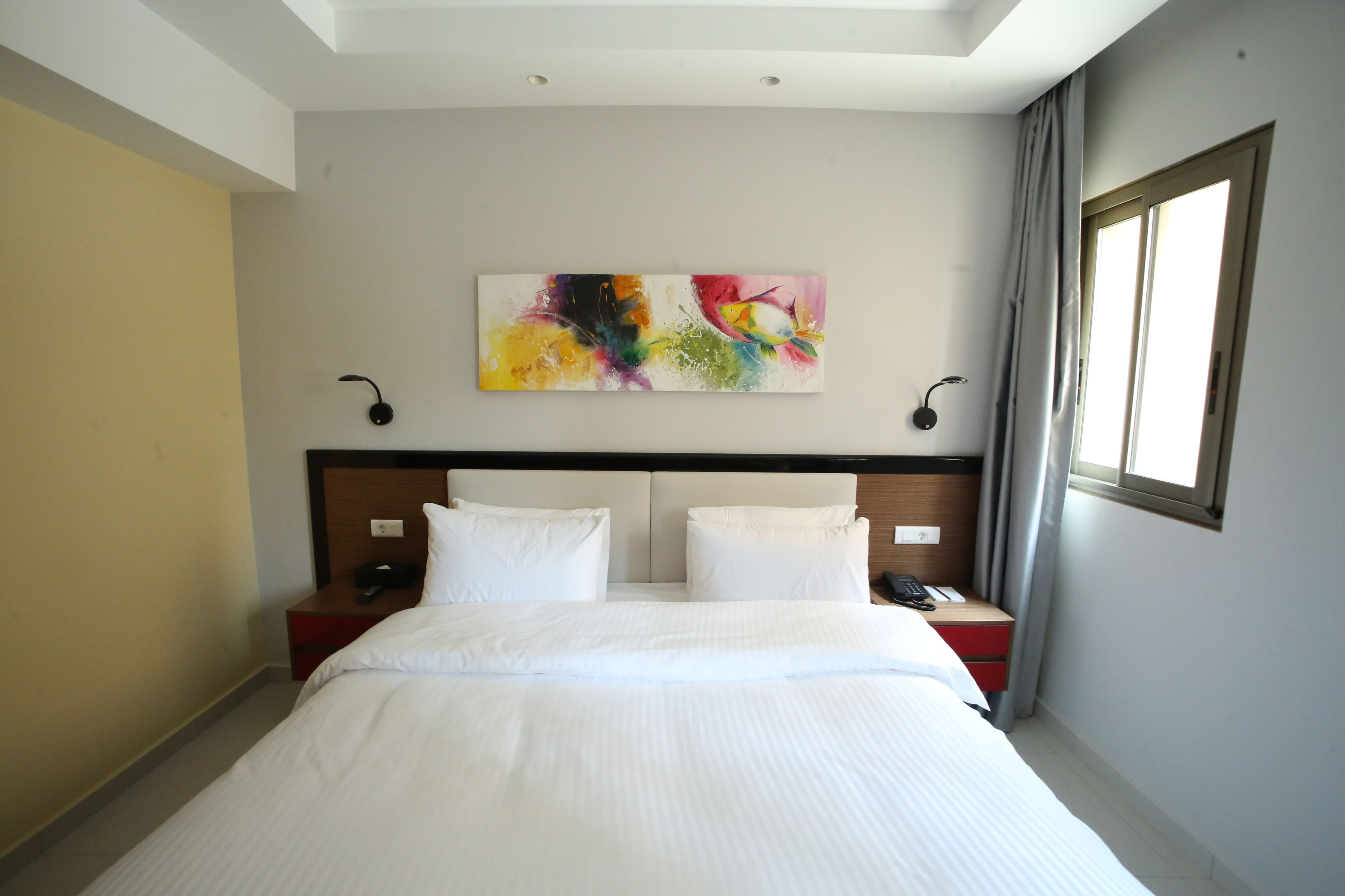 Quarto de hotel com uma cama grande, duas mesas de cabeceira, um quadro na parede e uma janela com cortinas.