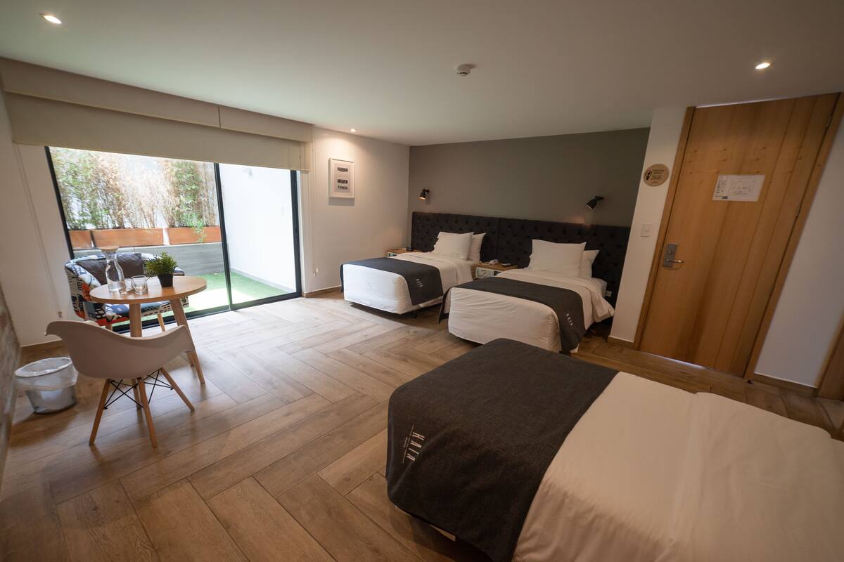 A modern hotel room with two beds, a dining area with a table and chairs, and a sliding glass door leading to an outdoor area.