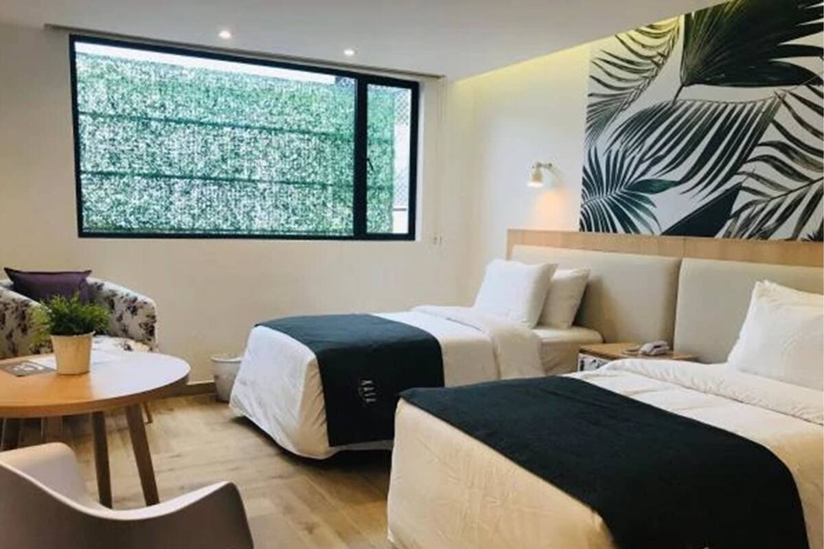 A hotel room with two beds, a small table, a chair, and a large window with a view of trees.