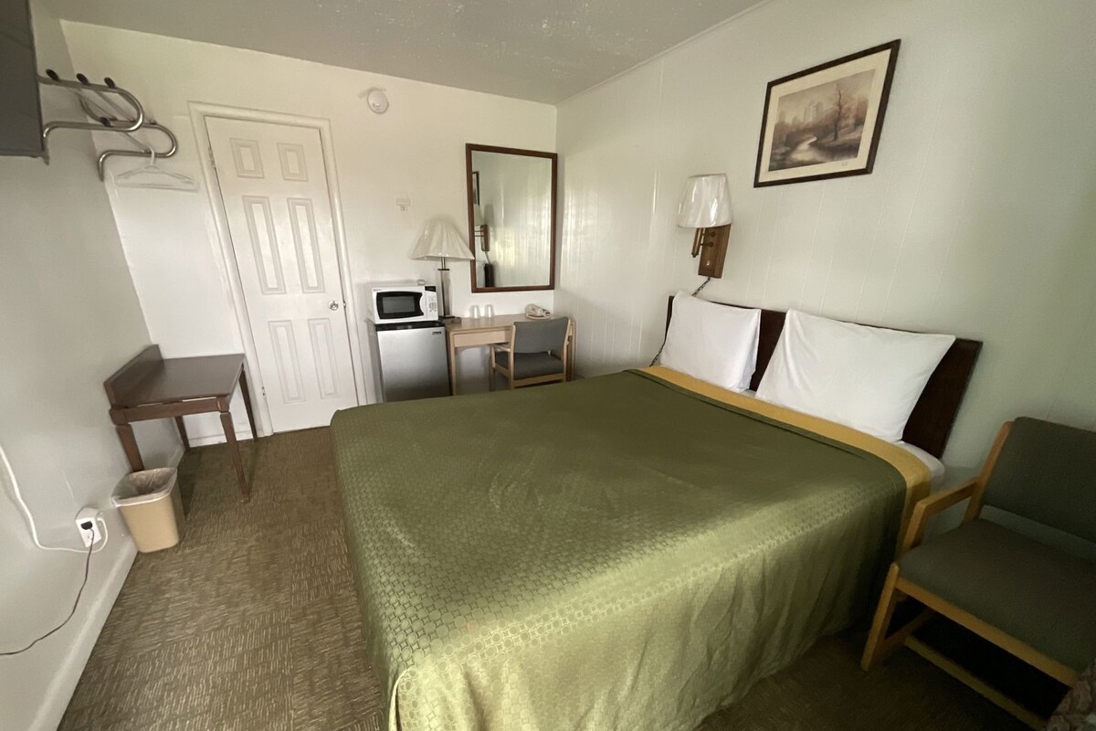 A hotel room with a bed, a desk, a chair, a microwave, a trash bin, and a picture on the wall.