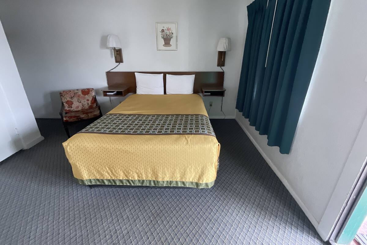 A hotel room with a bed, a chair, a nightstand, a lamp, a picture on the wall, and a window with curtains.