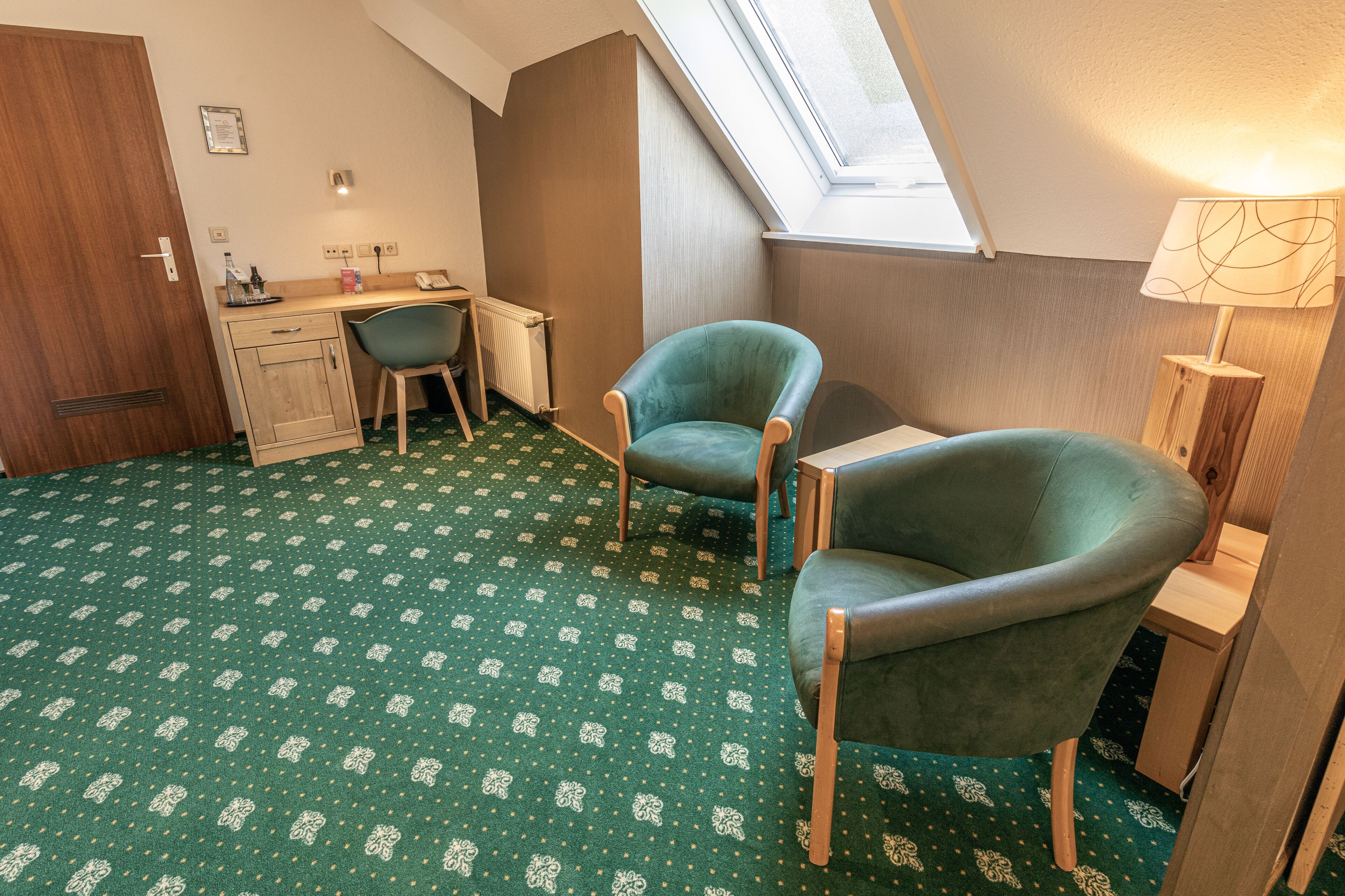 Een kleine, knusse kamer met een schuine kap, een bureau met een stoel, een tweede stoel, een radiator en een lamp.