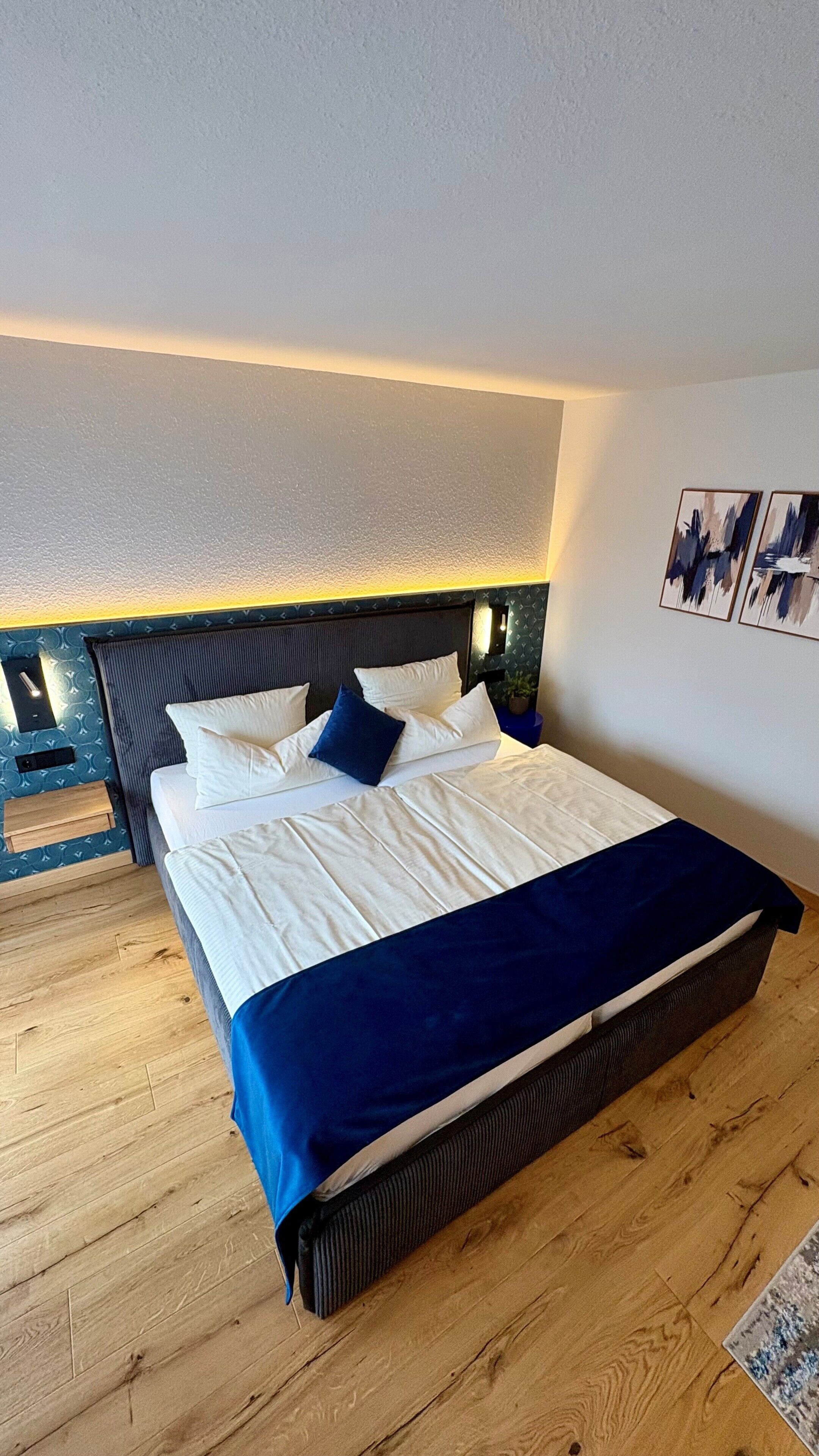 Une chambre à coucher comprenant un lit, un plancher en bois, une tête de lit, une table de chevet, des œuvres d’art murales et une petite table de nuit.