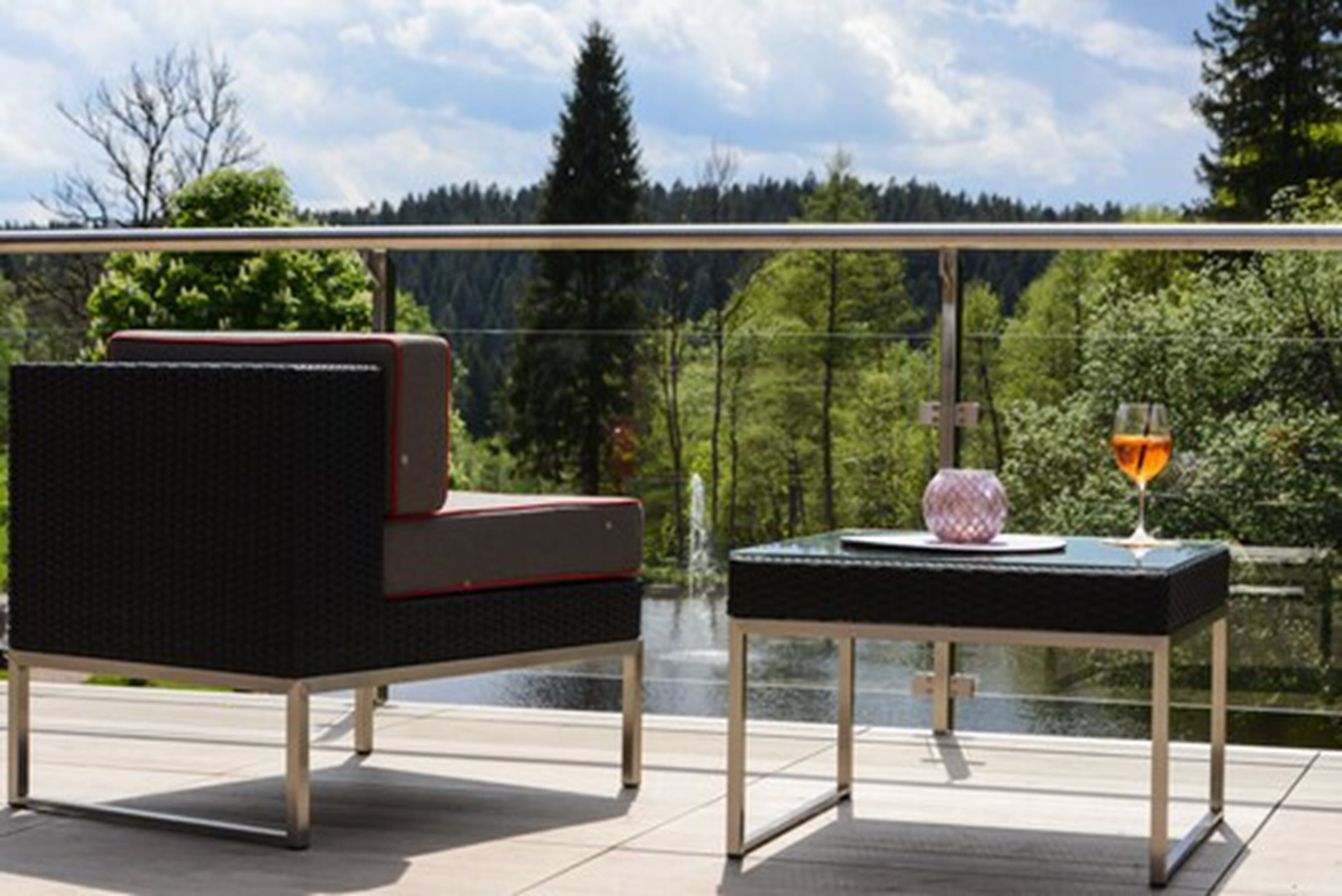 Un balcon avec un fauteuil en osier, une petite table sur laquelle se trouve un verre, et une vue sur une zone boisée.
