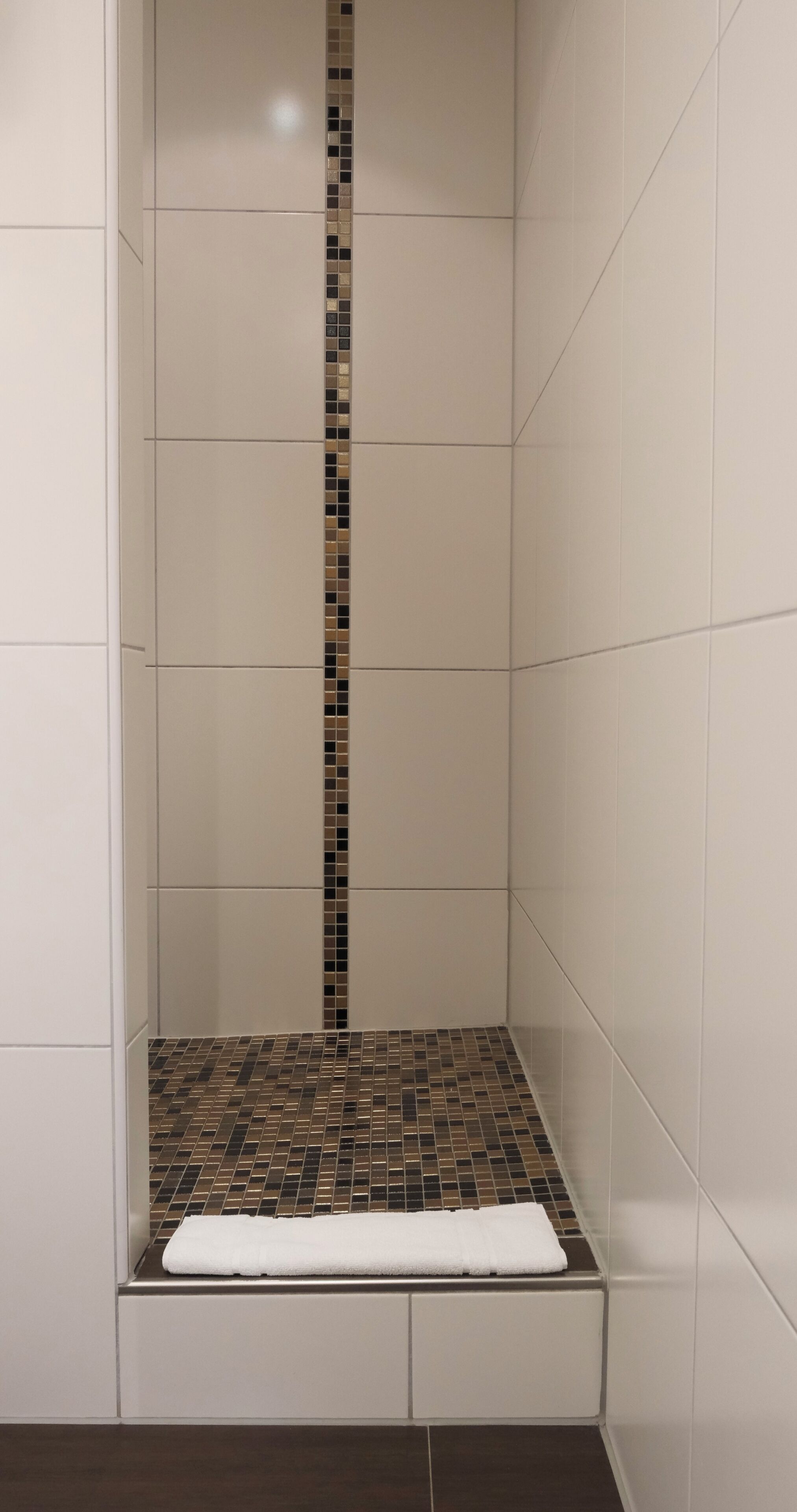 A shower with a tiled wall, a vertical strip of mosaic tiles, and a white towel on the floor.