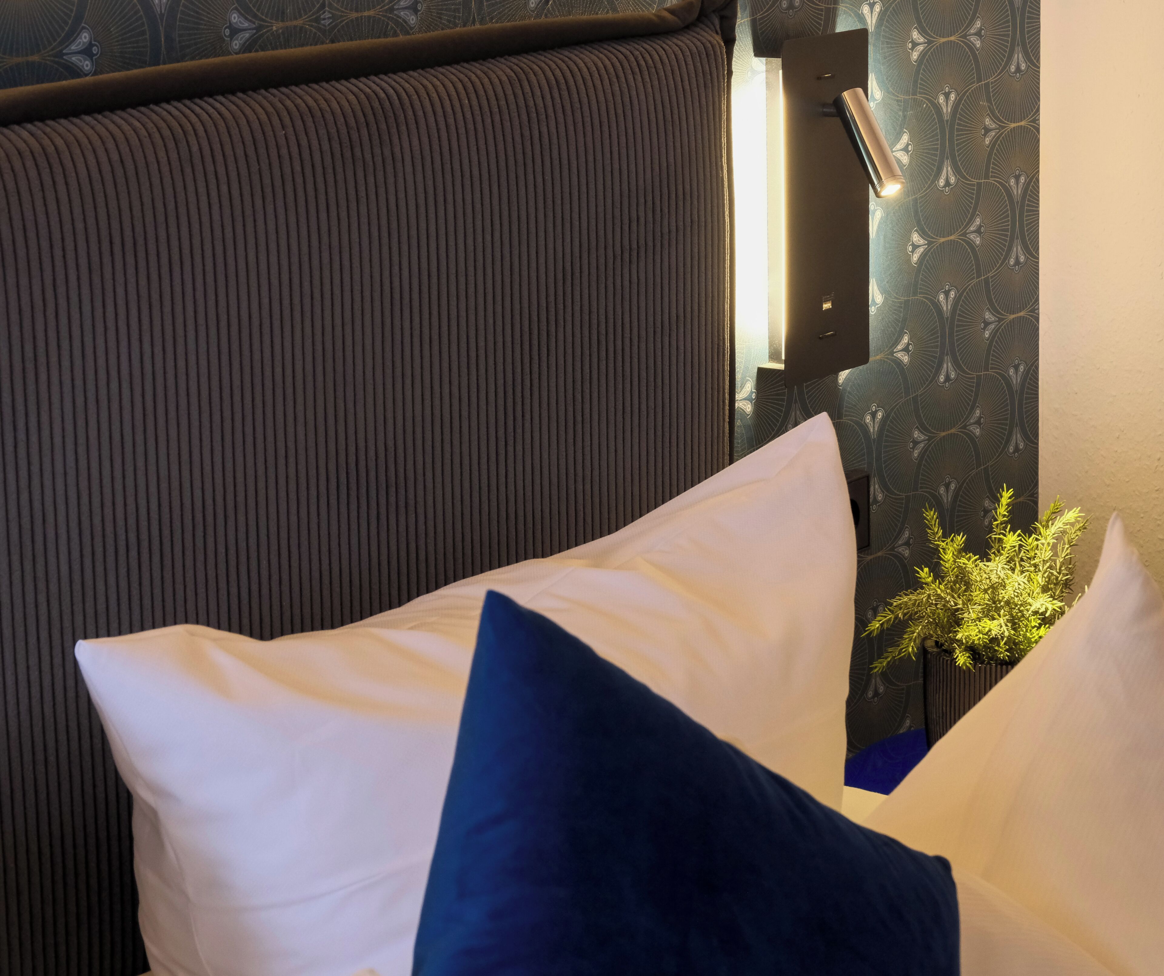 Una cama con almohadas blancas y azul oscuro, un cabecero oscuro y una pared con papel tapiz estampado.