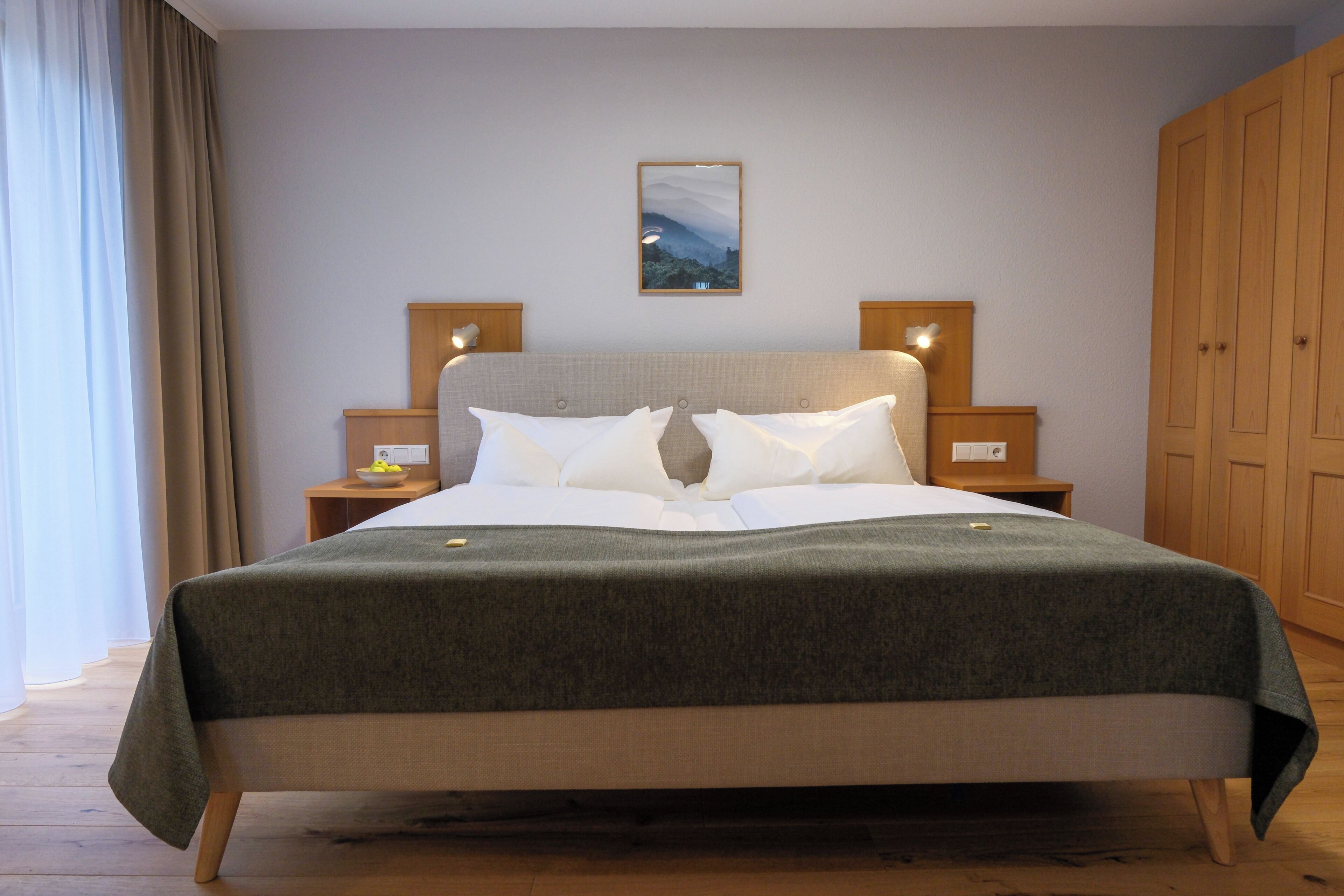 Una cama bien hecha con una colcha gris, dos mesitas de noche con lámparas, un cuadro en la pared y un armario de madera.