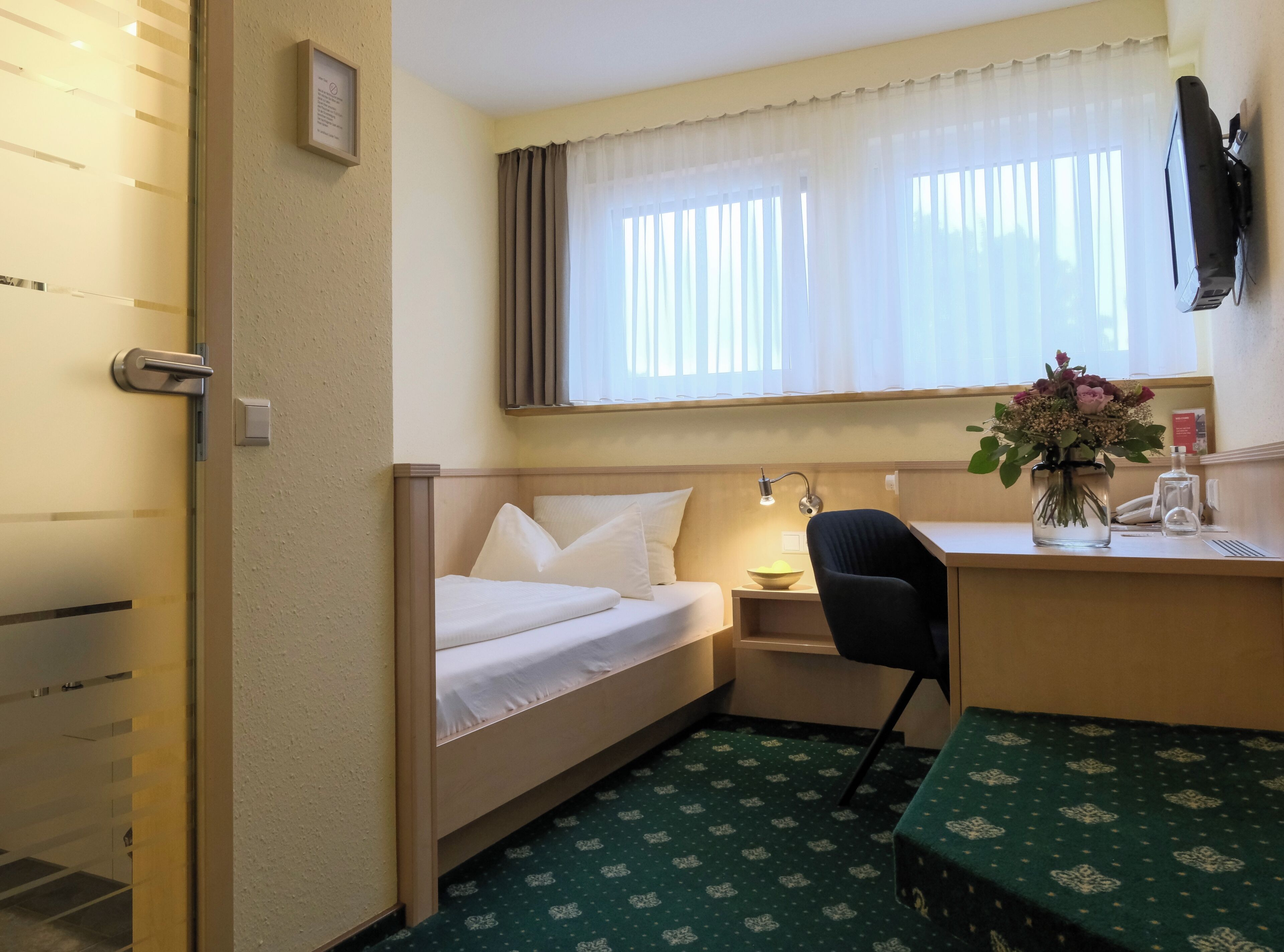 A compact hotel room with a single bed, a desk with a chair, a small table, a wall-mounted TV, and a window with sheer curtains.