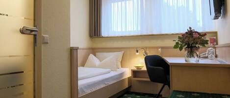 A compact hotel room with a single bed, a desk with a chair, a small table, a wall-mounted TV, and a window with sheer curtains.