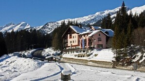 Estância de esqui alpina coberta de neve, com um edifício grande e varandas múltiplas, e um teleférico nas proximidades.