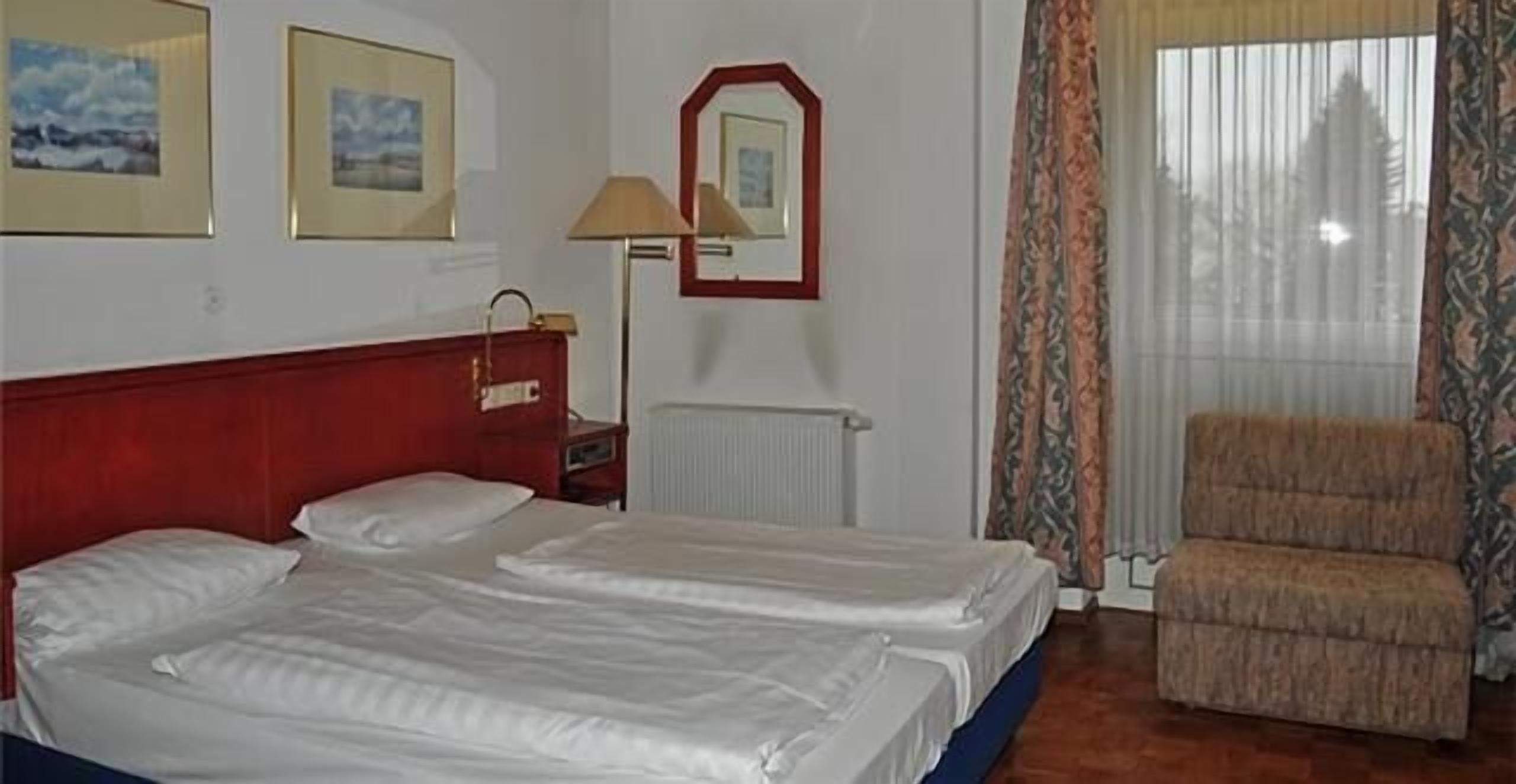 Une chambre d’hôtel avec deux lits, une tête de lit rouge, une chaise, un miroir et une fenêtre avec des rideaux.