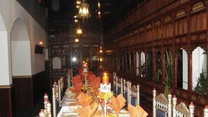 Un gran saló de banquets amb lluminàries ornamentals, una taula llarga coberta amb estesanes daurades i una paret amb finestres allargades i dibuixos.