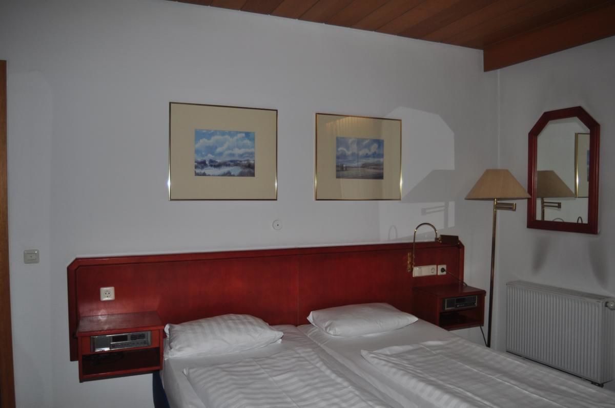 Quarto de hotel com cabeceira de madeira, duas camas, mesa de cabeceira, abajur, espelho e duas fotos emolduradas na parede.