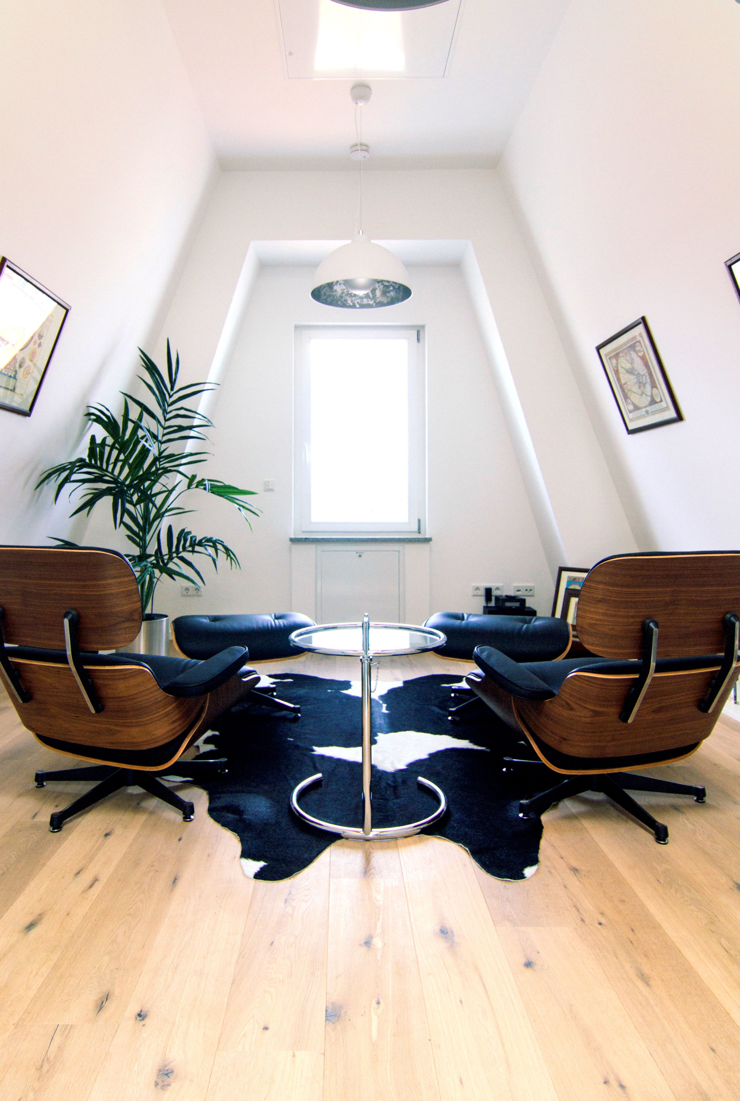 A room with a high ceiling, a large window, two wooden chairs with black cushions, a small round table, and a potted plant.
