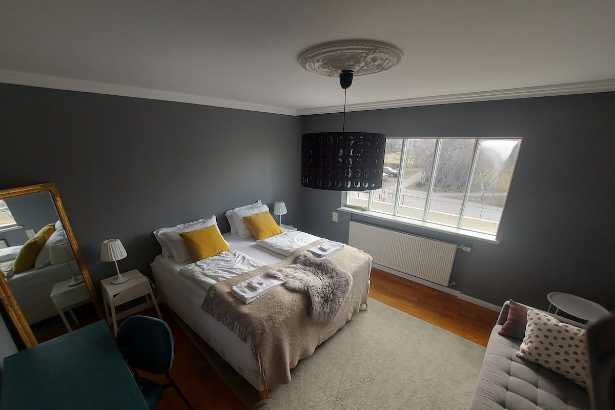 A bedroom with a bed, bedside tables, a desk, a chair, a mirror, a window, and a ceiling light fixture.