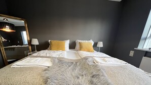Un letto rifatto con lenzuola bianche e due cuscini gialli, una soffice coperta grigia e un grande specchio a sinistra.