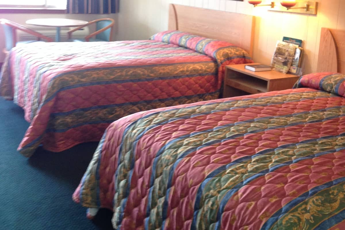A hotel room with two beds, a small table, and a chair. There is a window with curtains, a painting on the wall, and a lamp on the nightstand.