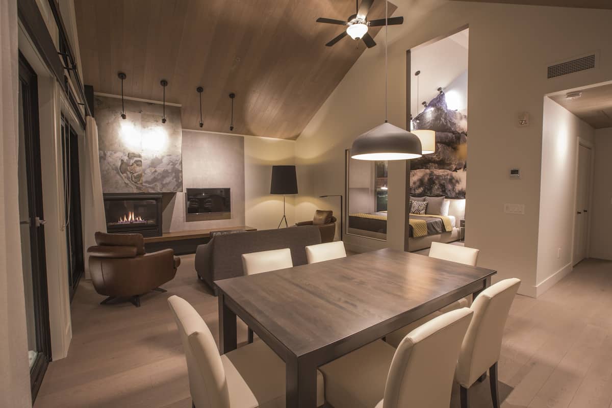 Un espace repas moderne avec une table et des chaises en bois, une cheminée et une vue sur un paysage montagneux, le tout reflété dans un grand miroir.