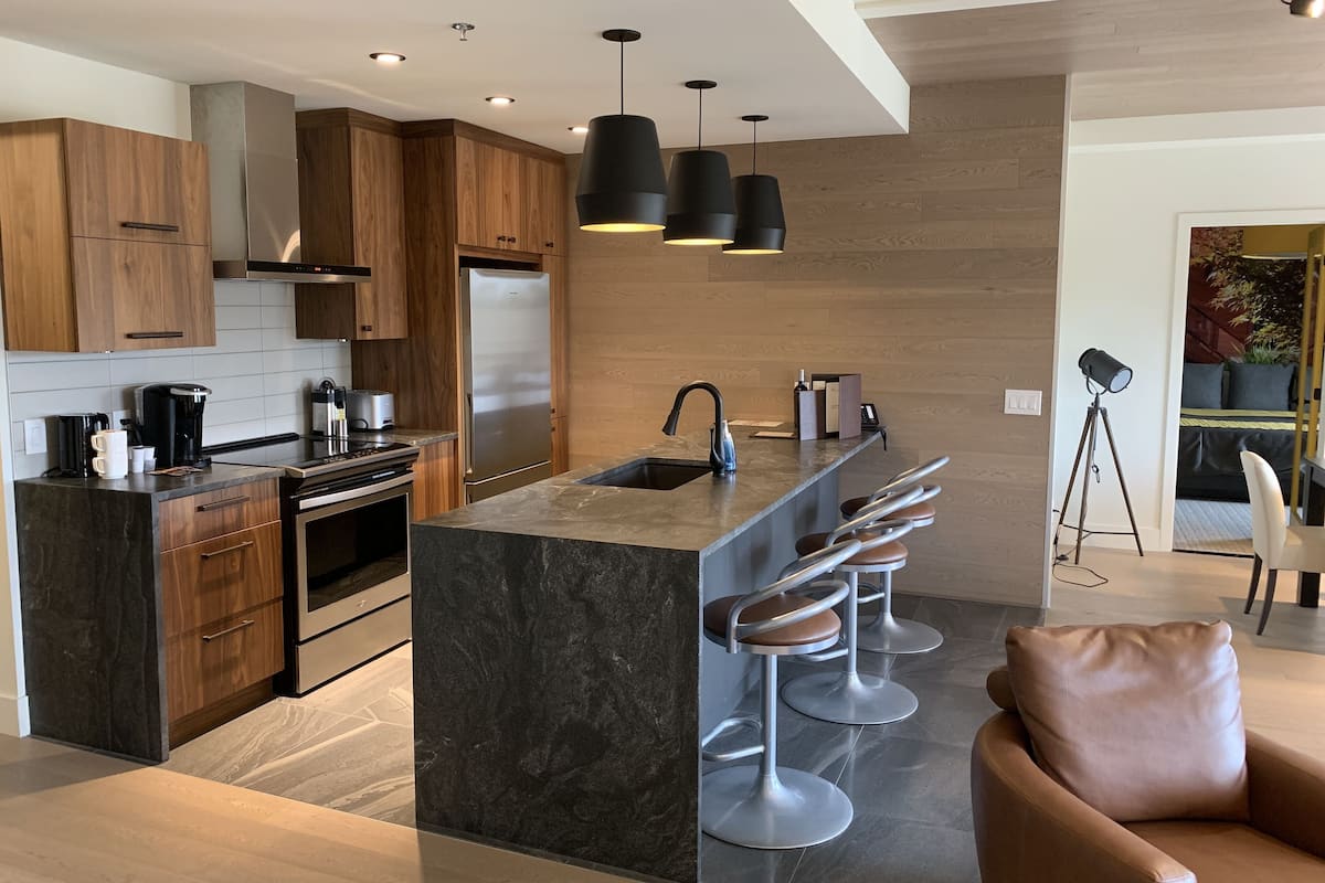 Une cuisine moderne avec des meubles en bois, un réfrigérateur en acier inoxydable, un four encastré, une machine à café et un coin bar avec des tabourets.