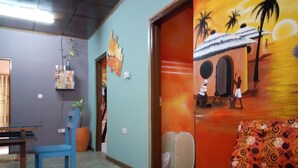 A room with a blue chair, a sofa with a patterned cover, and a painting of a tropical scene on the wall. - Feehi's Place - Hostel (Accra)
