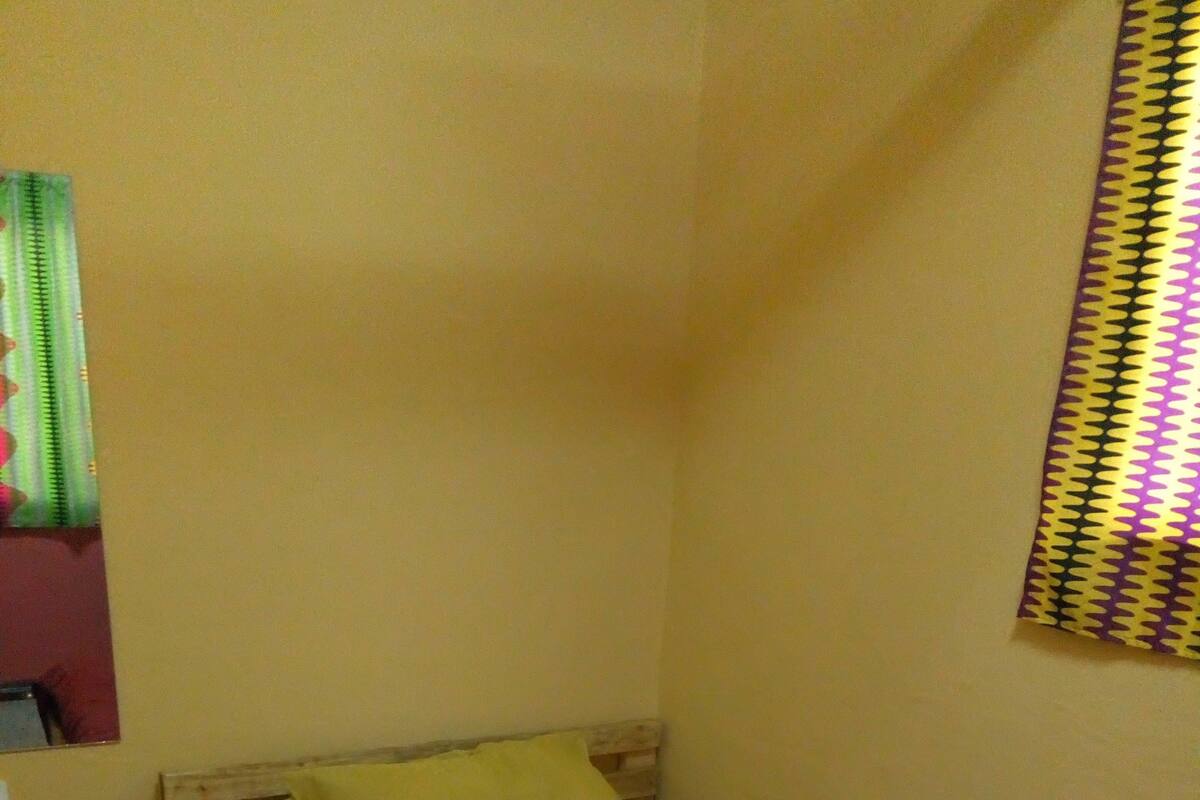 Ein kleines Zimmer mit einem Einzelbett, einem Holz-Kopfteil, einem gelben Kopfkissen und einer roten Decke. Ein kleiner Nachttisch mit Lampe und Steckdose ist vorhanden.
