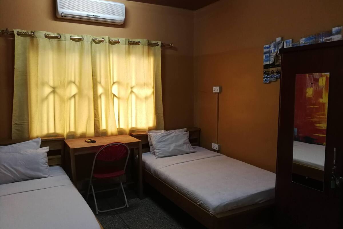 Ein kleines Zimmer mit einem Bett, einem Schreibtisch und einem Stuhl. Es gibt ein Fenster mit Vorhängen, eine Klimaanlage und eine Wandsteckdose.