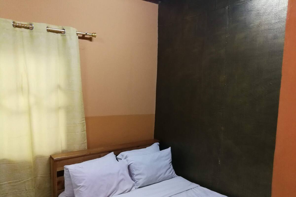 Ein kleines Zimmer mit einem Einzelbett, einem Holzkopfstück, weißer Bettwäsche und einem halb geöffneten Vorhang.