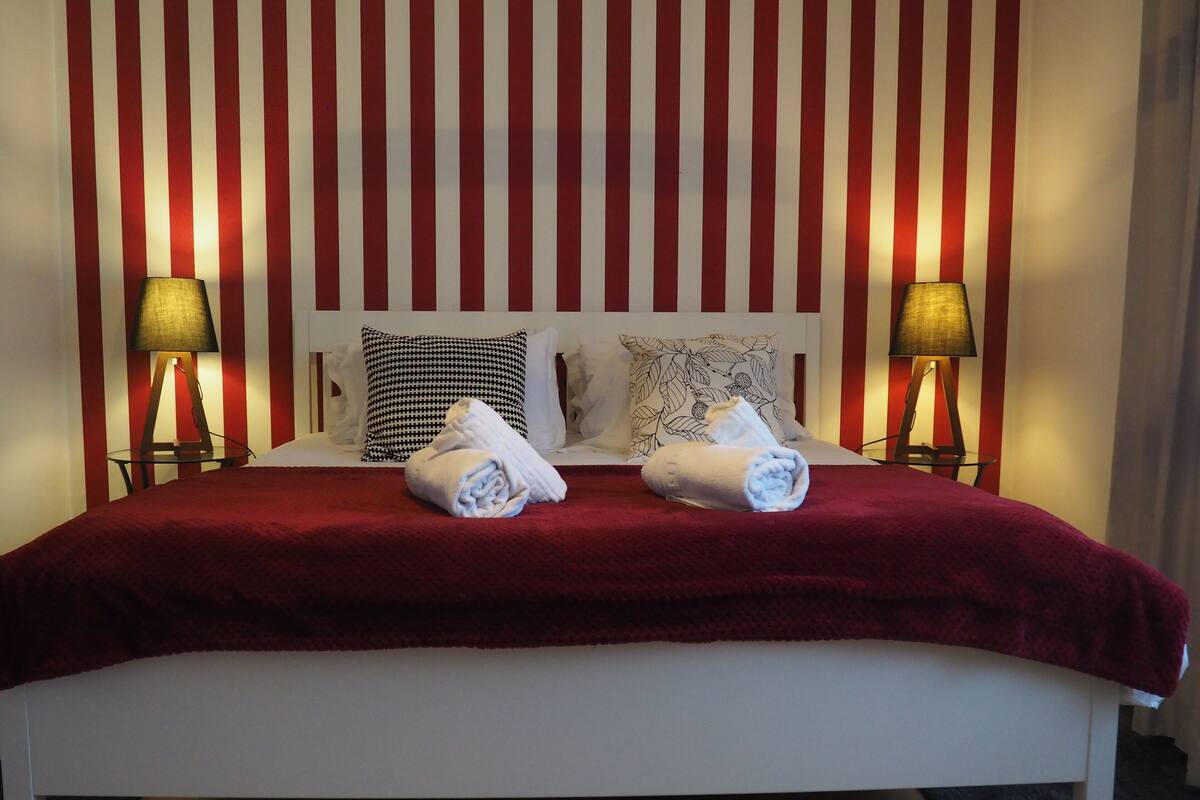 A bed with a red blanket, two pillows, and two rolled towels. There are two bedside tables with lamps.