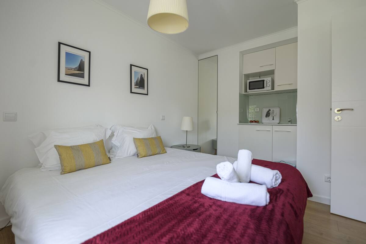 A neatly made bed with white and yellow pillows, a red blanket, and rolled towels. There is a white wardrobe with a microwave above it.
