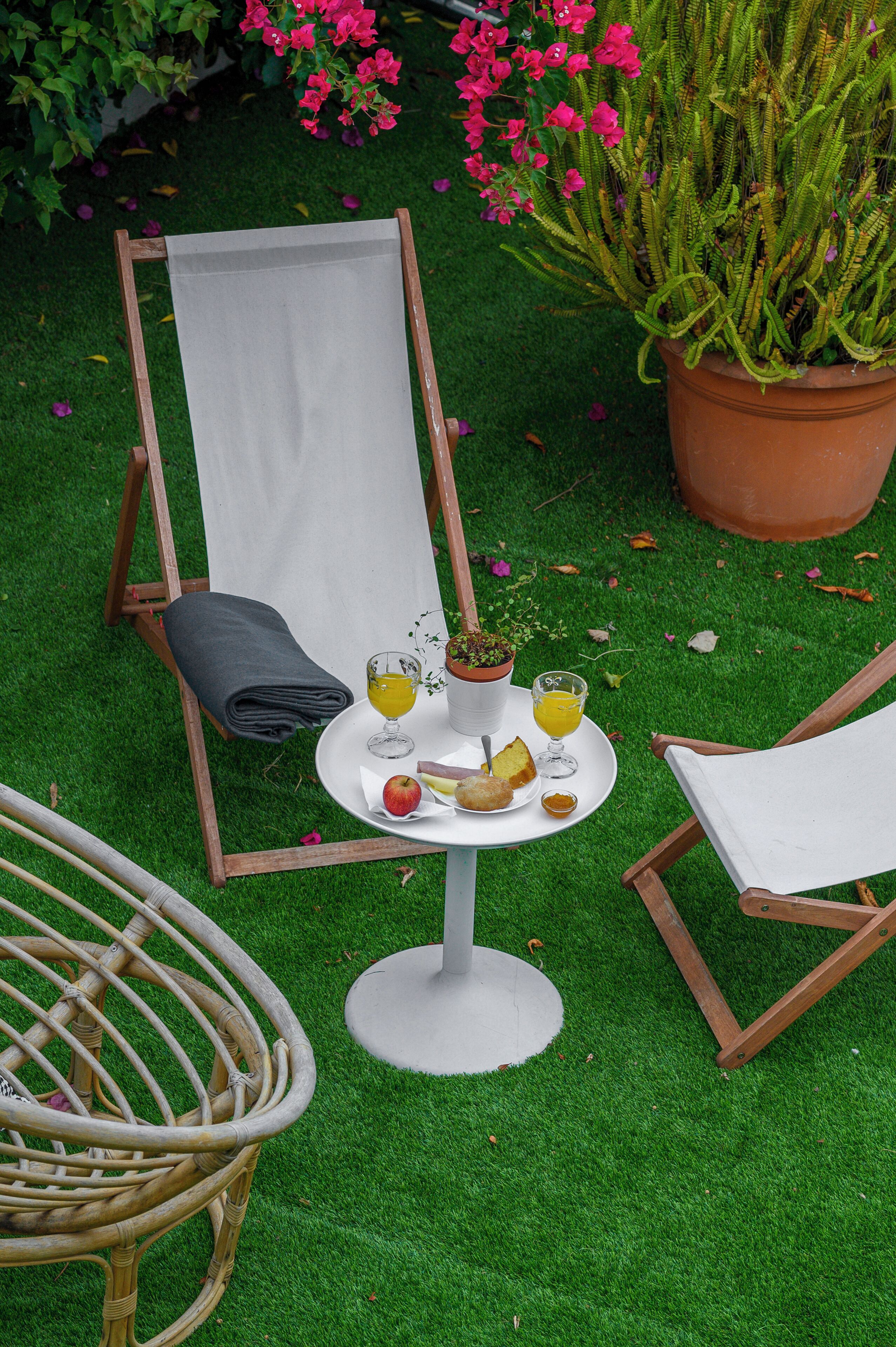 Un jardín con una silla de descanso, una mesita con bebidas y aperitivos, y una planta en maceta.