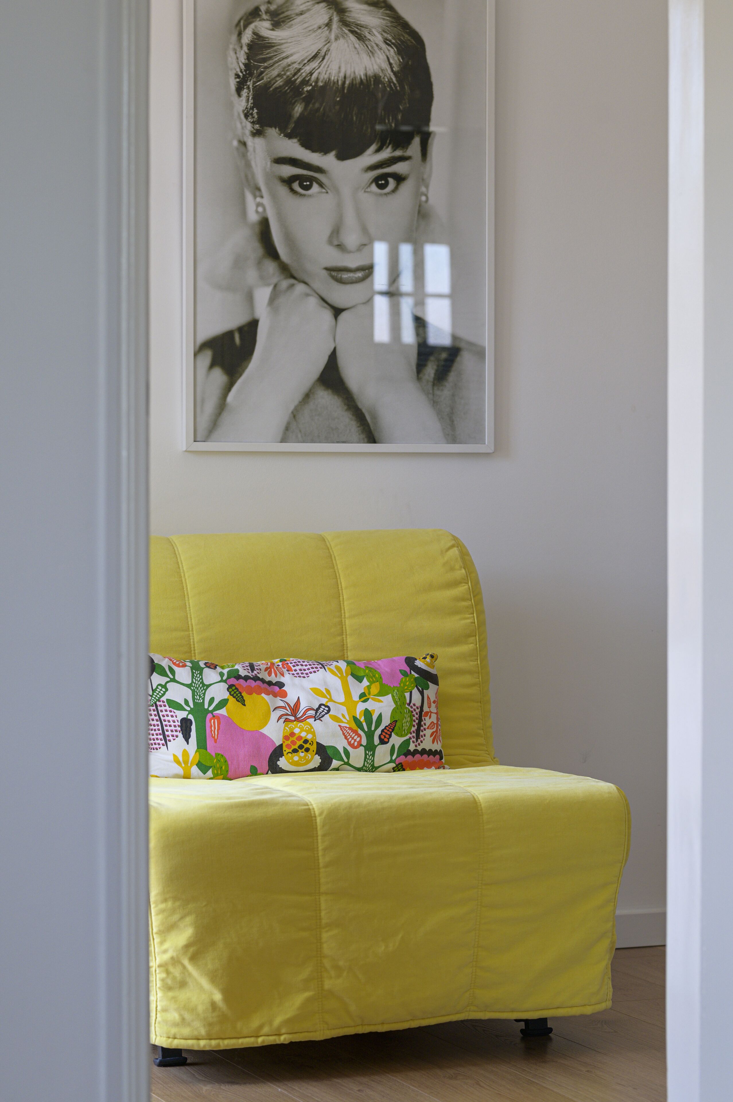 Un sillón amarillo con un cojín floral de colores, ubicado en una habitación con una fotografía en blanco y negro enmarcada en la pared.
