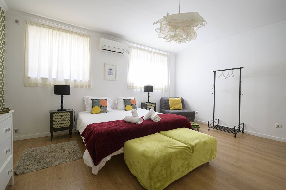 A bedroom with a bed, bedside tables, a desk, a chair, a hanging light fixture, and a wardrobe.