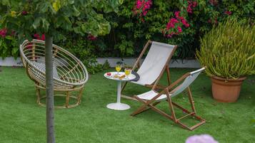 Relax in a serene outdoor setting with a wicker chair and lounge chair surrounded by lush greenery and flowering plants.