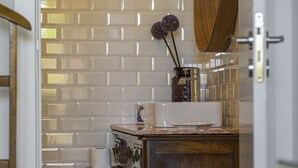 A bathroom with a white tiled wall, a wooden vanity, a mirror, and a trash bin. - Casa das Hortênsias - Charming Guest House (Sintra)