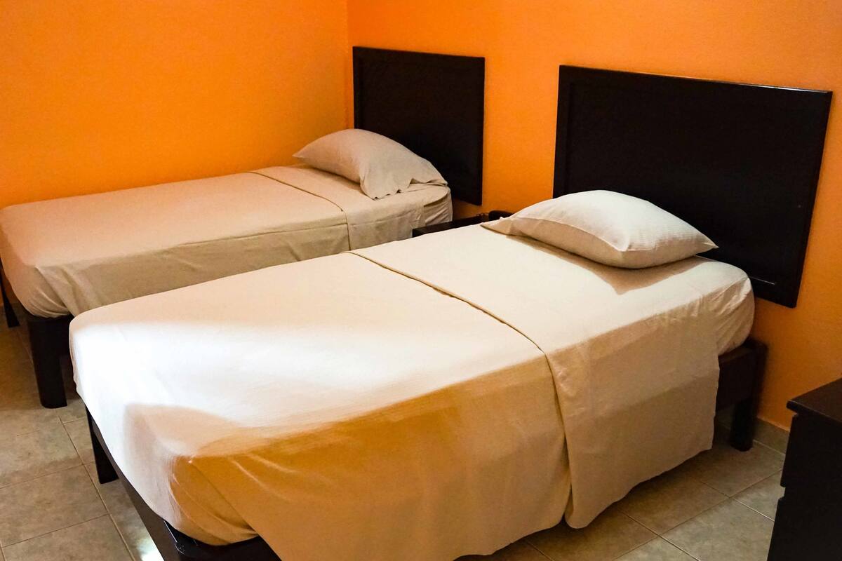Een kamer met twee eenpersoonsbedden, elk met een kussen en hoofdbord. De muren zijn oranje en de vloer is betegeld.