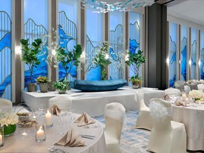 Un salón de banquetes con mesas redondas, sillas blancas y un banco acolchado azul.