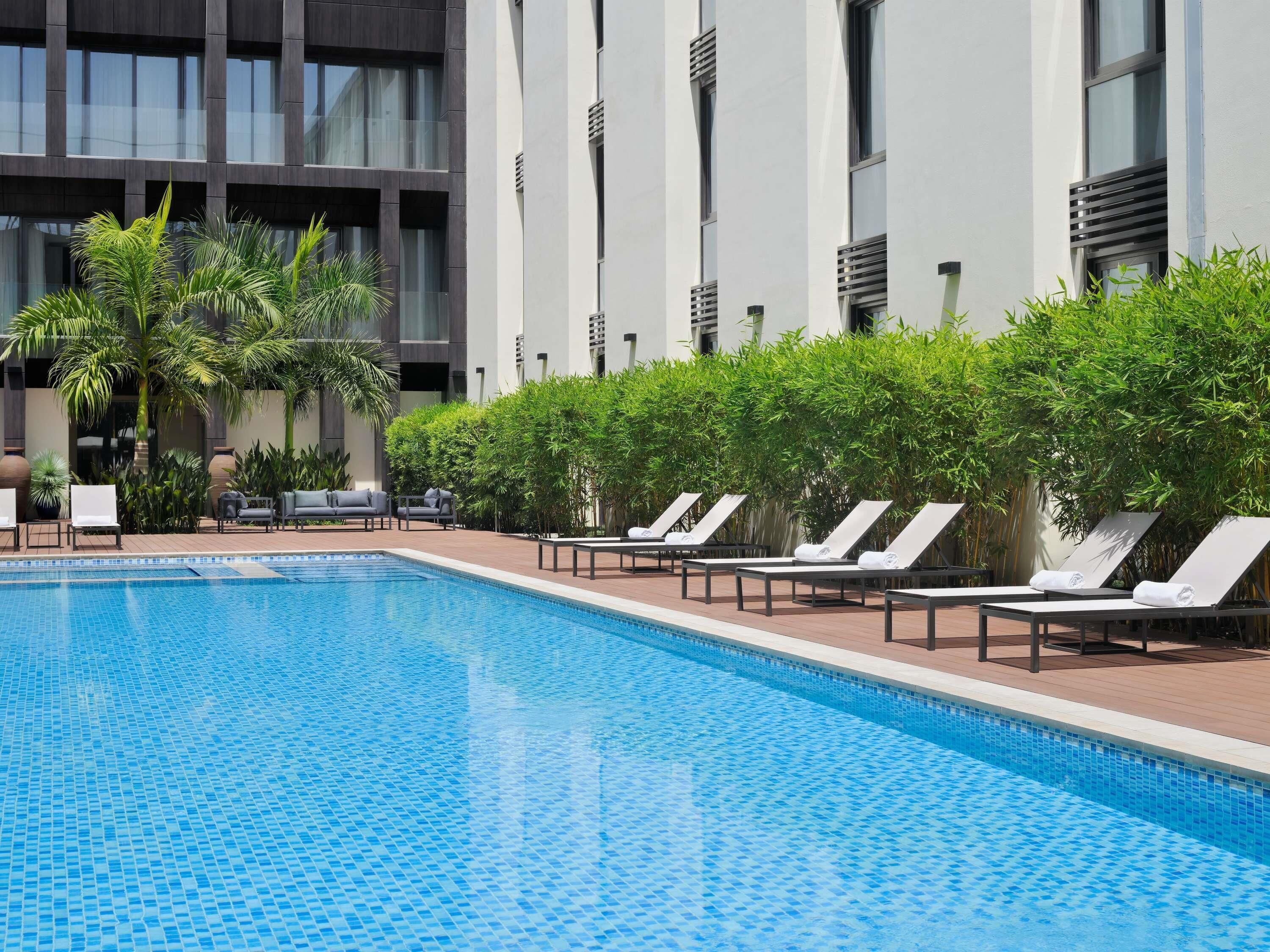 Una piscina con sillones de descanso, rodeada de follaje y edificios modernos.