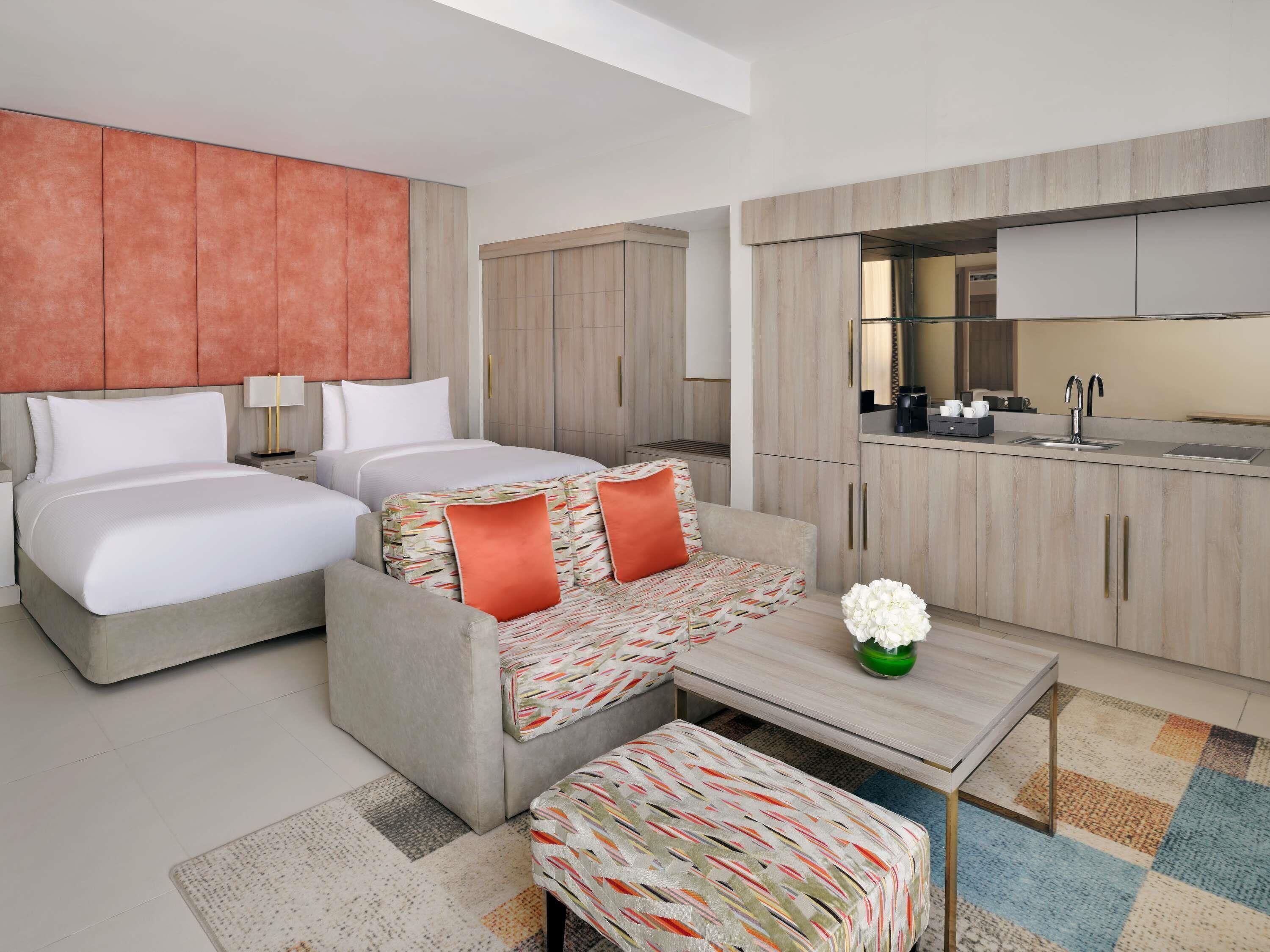 A modern hotel room with two beds, a sofa, a small table, and a kitchenette.